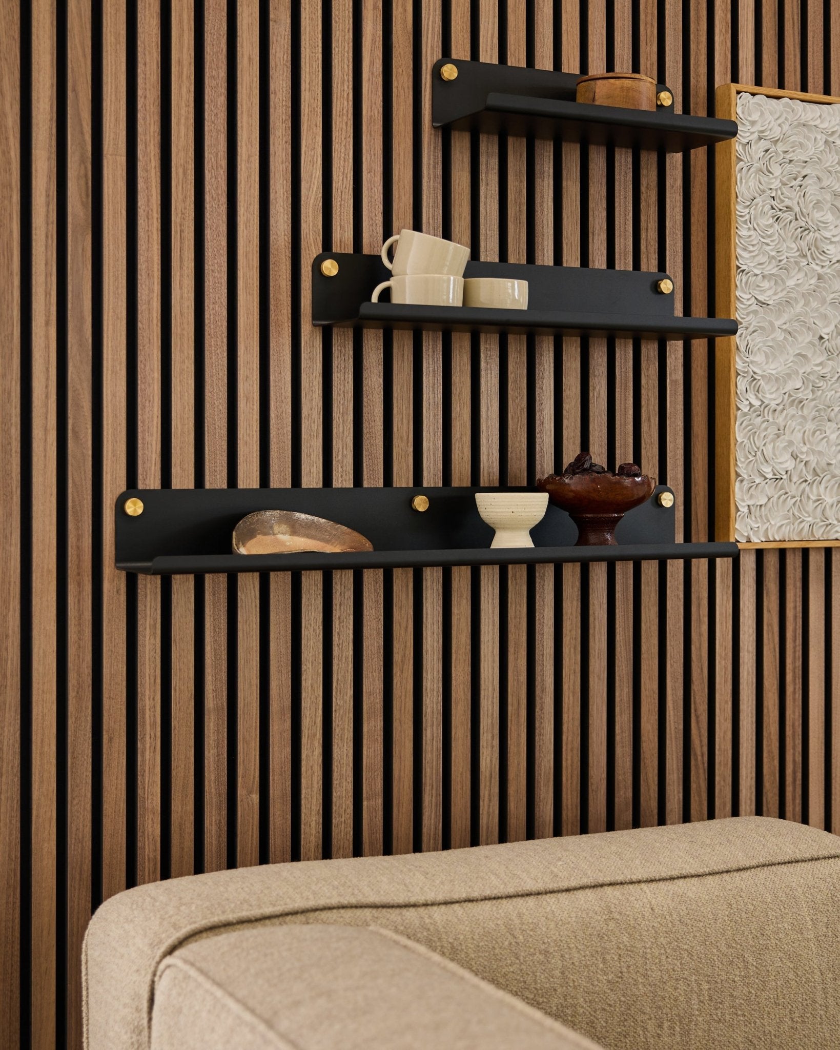 Trois étagères black, dont l’étagère large WoodUpp Norr (75,9 cm), présentent des objets déco sur un mur en Panneau acoustique à lattes de bois au-dessus d’un canapé beige ; une partie de l’œuvre est visible.